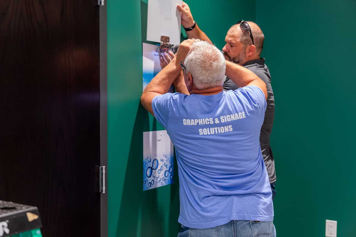 P-R-A-C-T-I-C-E Core Values with BPI Color: Priority 3 Close-up view of two men installing custom graphics and images for CFR's History Wall.