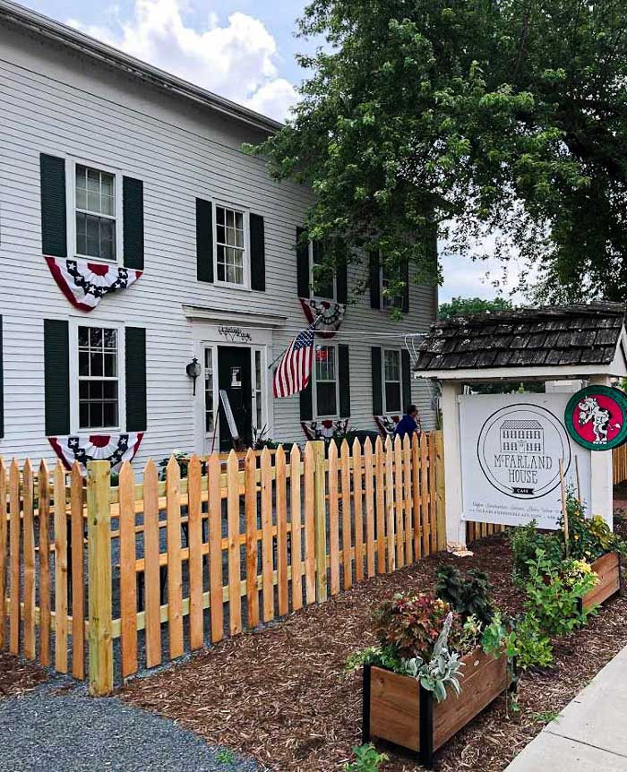 McFarland House Cafe 4 An outdoor sign displaying the McFarland House Logo with a white building to the left of the sign and wooden picket fence.