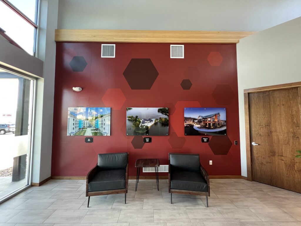HBI lobby wall display featuring large mounted project photos on a red wall with hexagon graphics and two black chairs below.