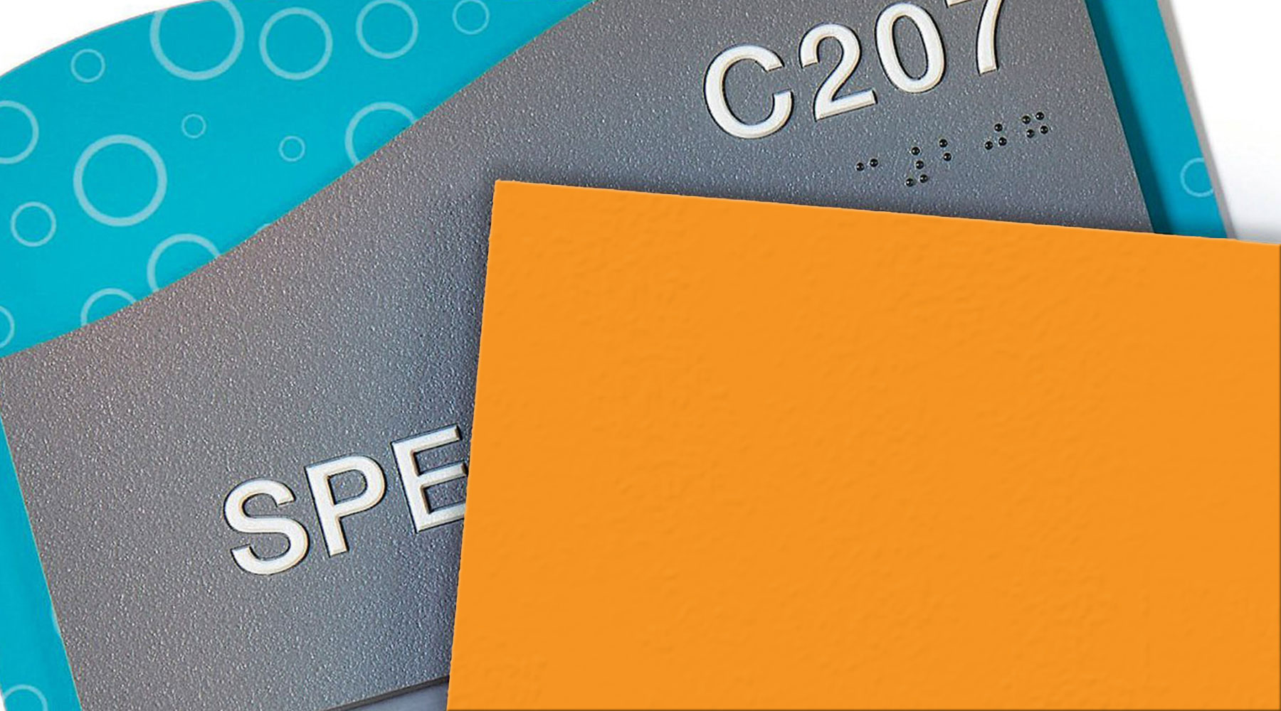 ADA Signage featuring a textured gray sign with raised white lettering and Braille, accented by a blue pattern and partially covered by an orange color swatch.