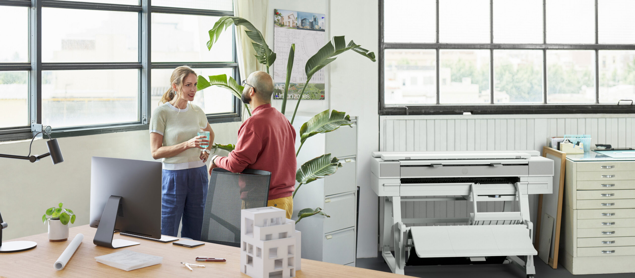 Printing Solutions Menu 4 HP Wide Format Printers – Two professionals talking in a bright office space next to an HP wide format printer, surrounded by architectural tools and modern furniture.