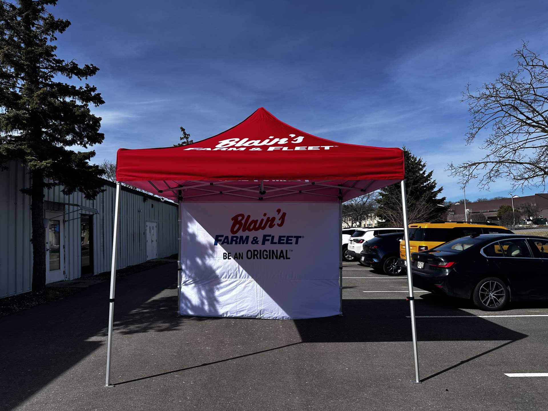 Tradeshow Event Tent with a red canopy and branded white back panel reading "Blain's Farm & Fleet – Be An Original™" set up in a parking lot outside a metal building.