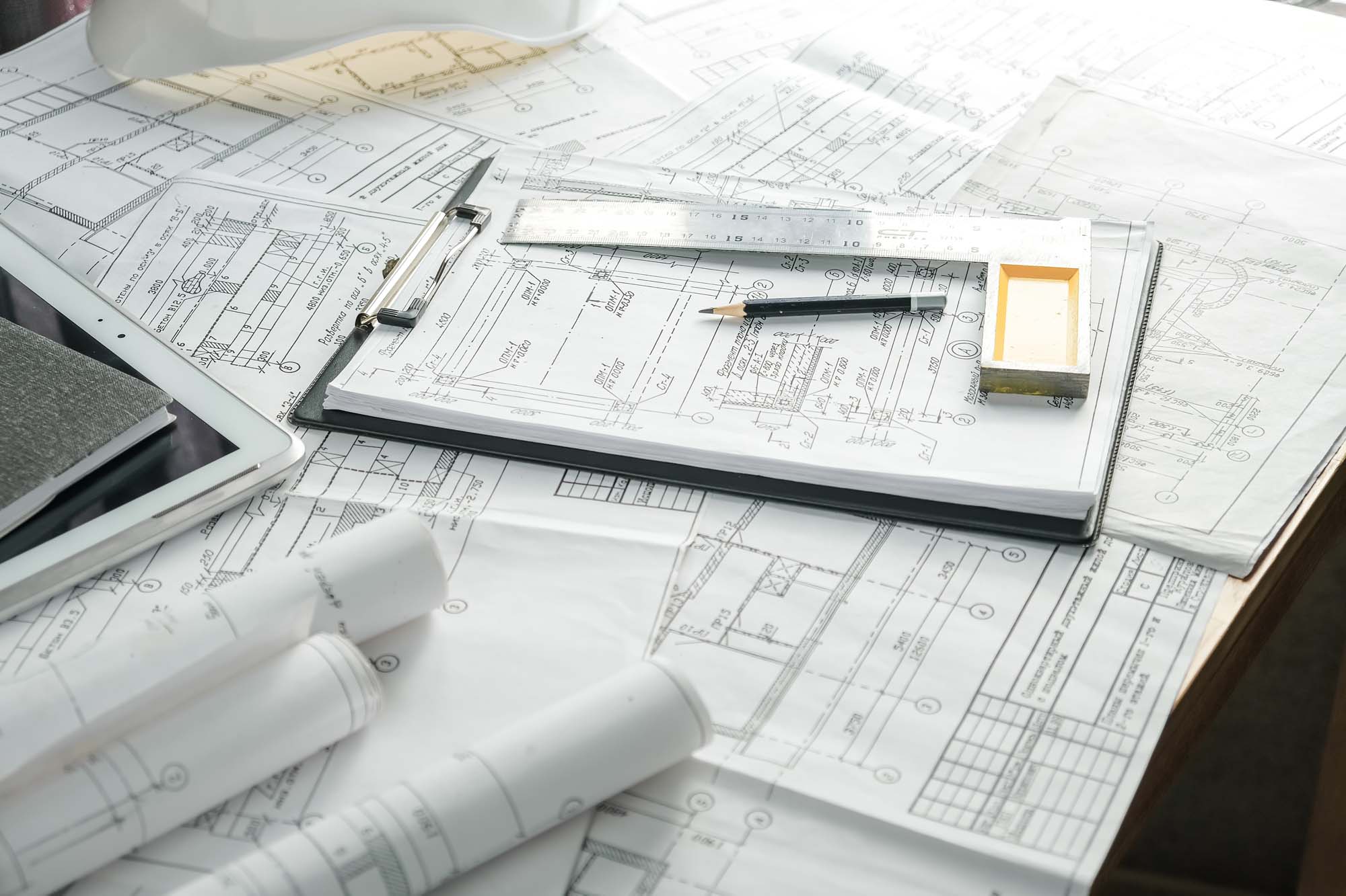 Professional Services Menu 3 Close-Out Docs – A tabletop covered with architectural drawings, blueprints, a clipboard, ruler, pencil, and tablet, representing construction project documentation.