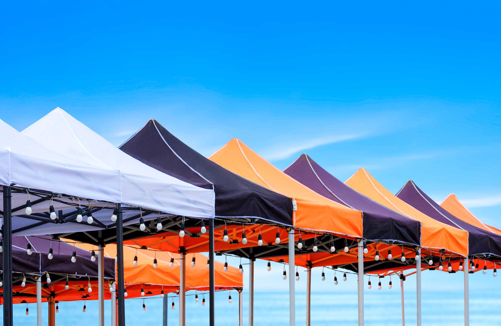 Tradeshow Event Tent setup with multiple colorful canopy tents in white, black, orange, and purple, lined up along a waterfront with hanging string lights.