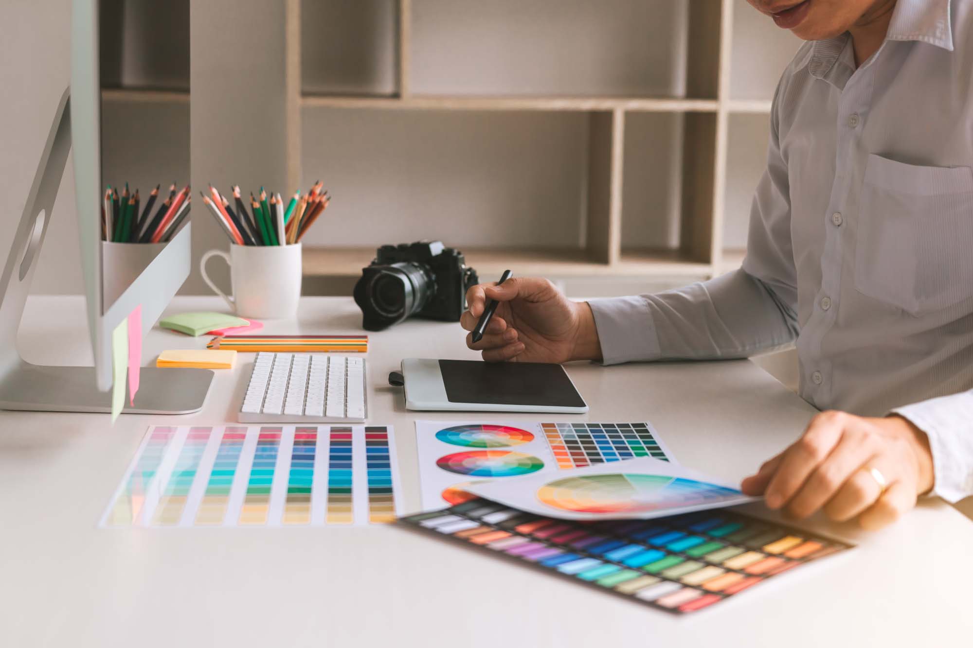 Creative Studio Menu 2 Graphic Design – A designer working at a modern desk with a digital tablet, color swatches, and a desktop computer, reviewing color palettes and design tools.