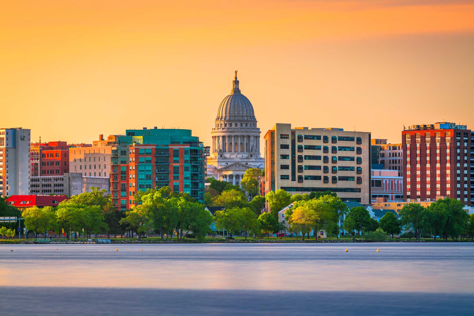 Madison East: Sunset view of downtown Madison, Wisconsin, featuring the Wisconsin State Capitol dome framed by modern buildings and trees along the lakefront.