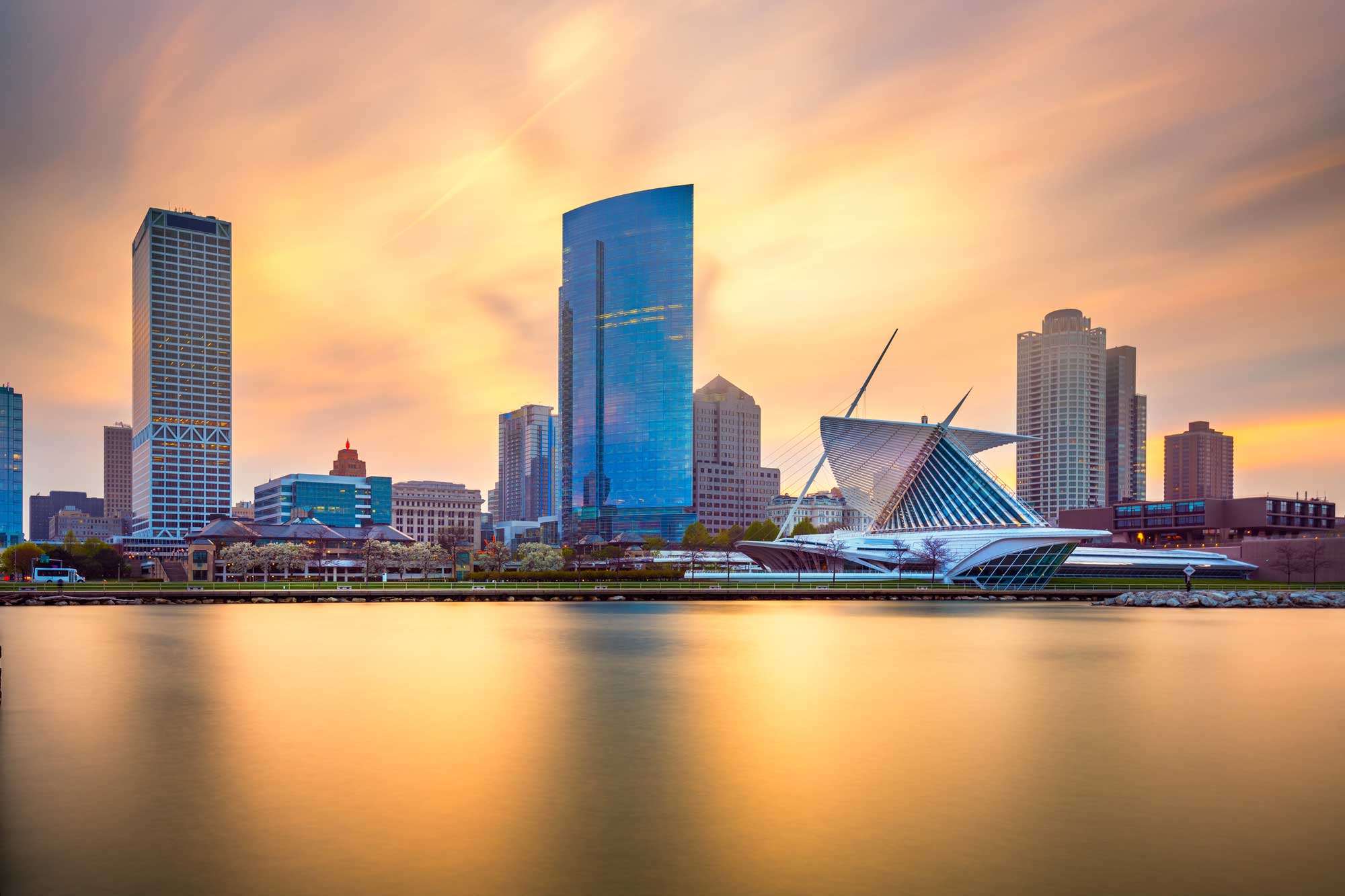 Scenic view of the Milwaukee skyline at sunset, featuring the Milwaukee Art Museum and high-rise buildings reflected in calm lake waters.