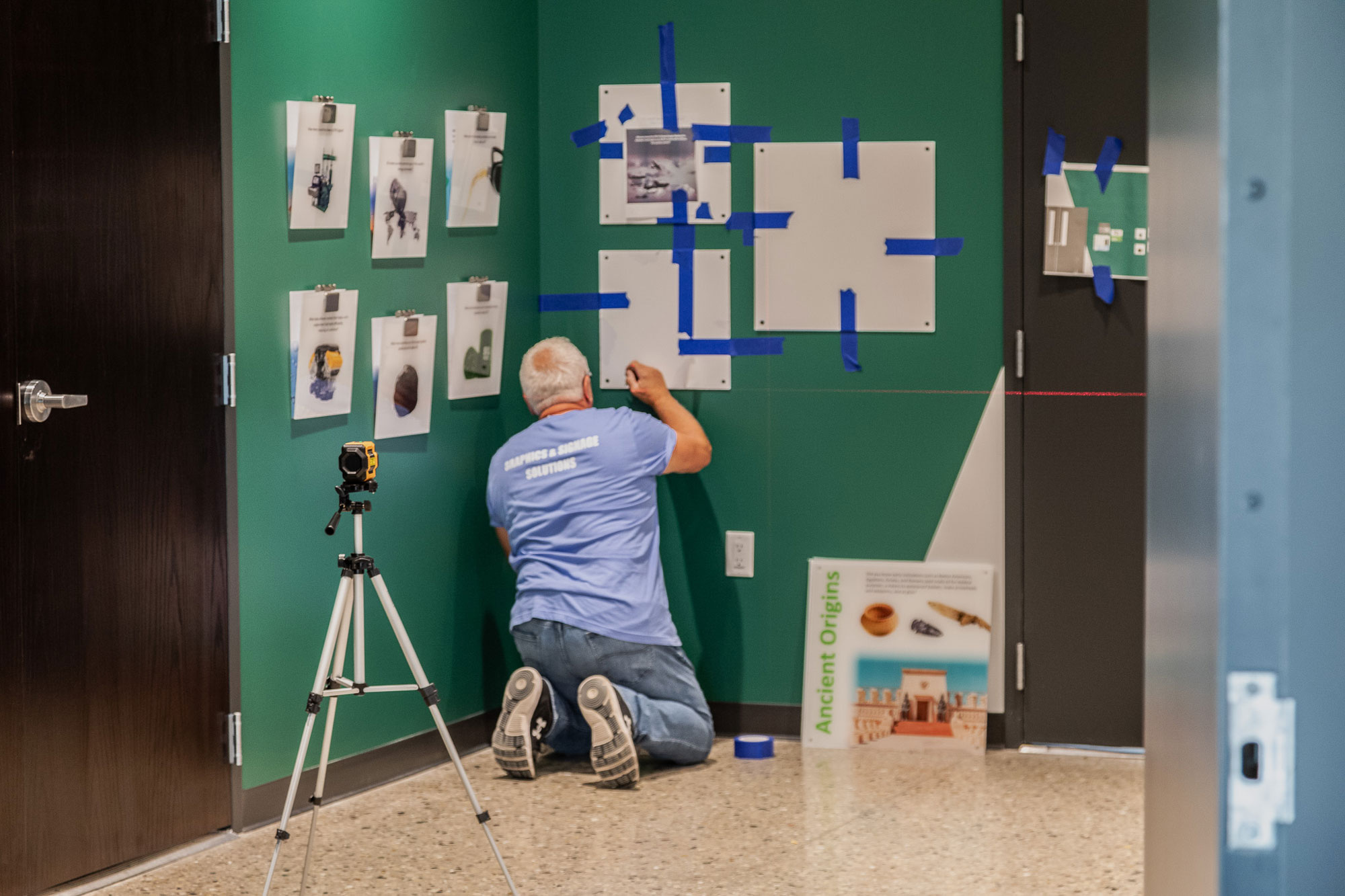 Installation for CFR Engines' Graphics: Technician kneeling while aligning and taping wall graphics for CFR Engines, using a laser level and tripod for precision installation.