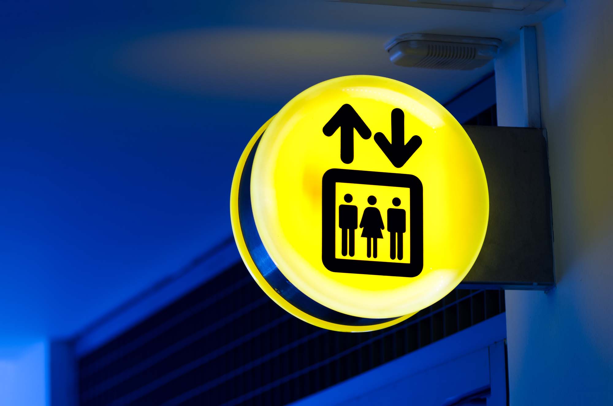 Illuminated Signage: Bright yellow circular illuminated elevator sign featuring up and down arrows and pictograms of three people, mounted on an interior wall with blue lighting.
