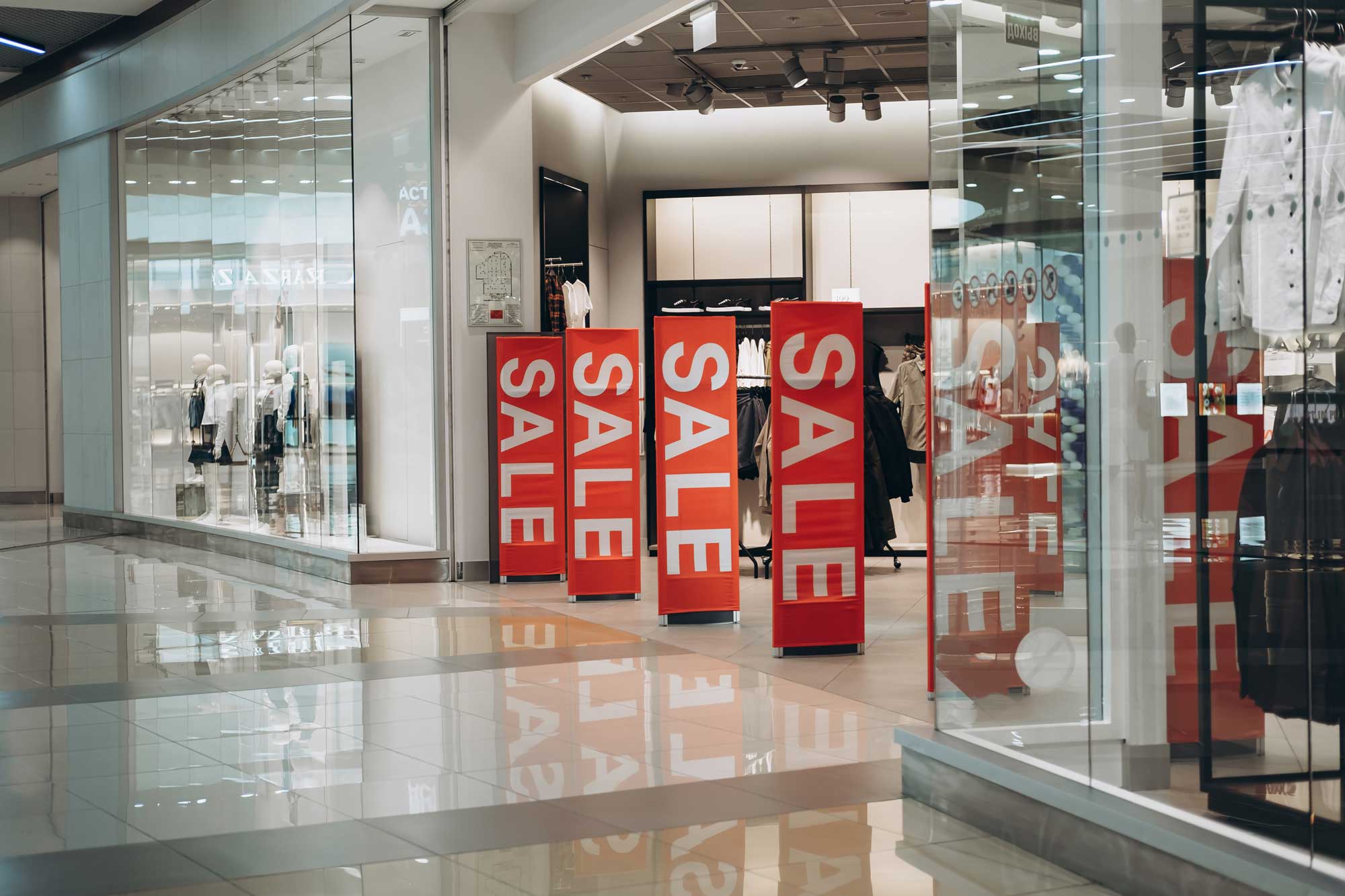 Stand Up Banners with bold red SALE lettering displayed at the entrance of a retail clothing store inside a shopping mall.