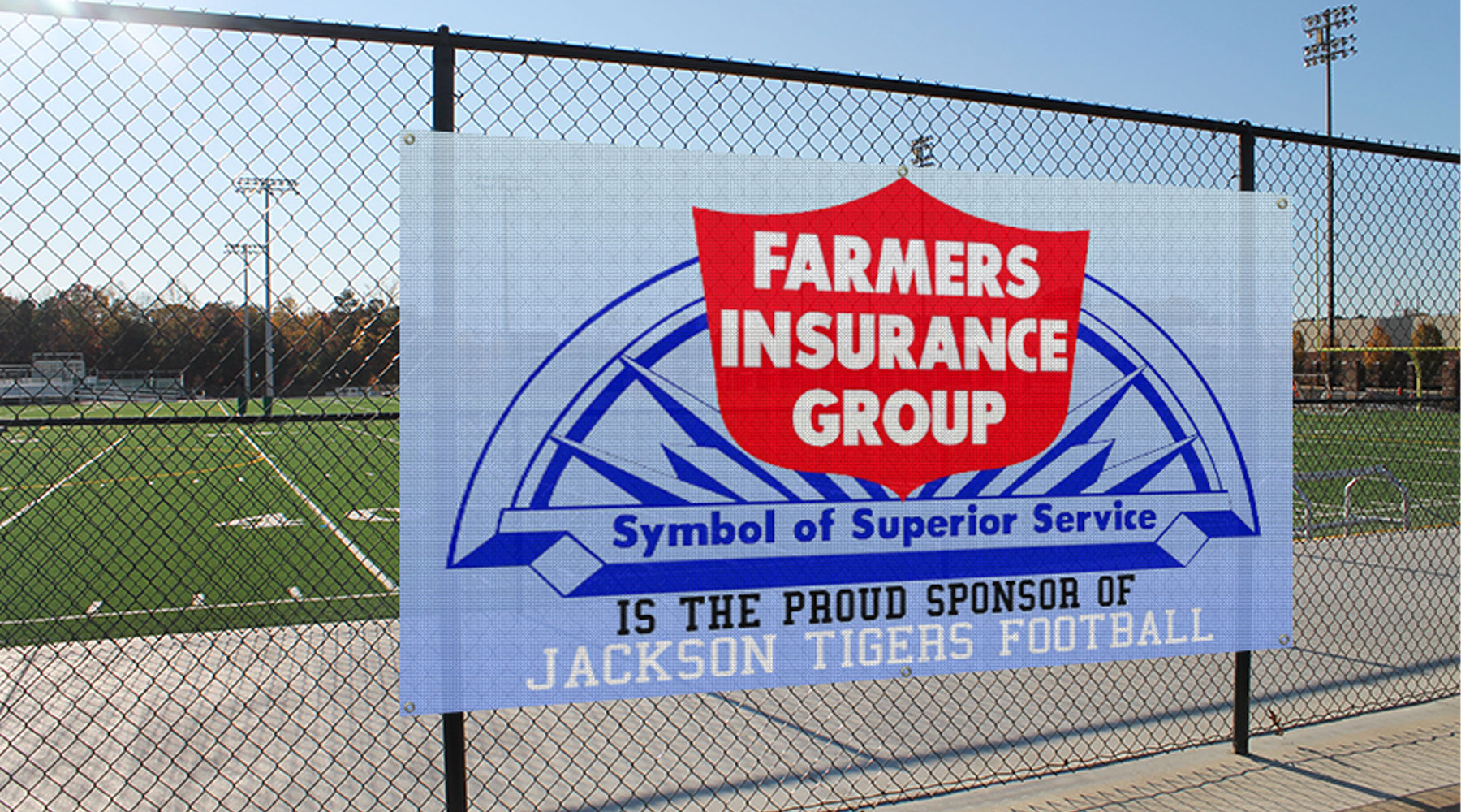 Mesh Hanging Banner for Farmers Insurance Group attached to a chain-link fence at a football field, displaying the company logo and the message “Symbol of Superior Service” and “Proud Sponsor of Jackson Tigers Football.”
