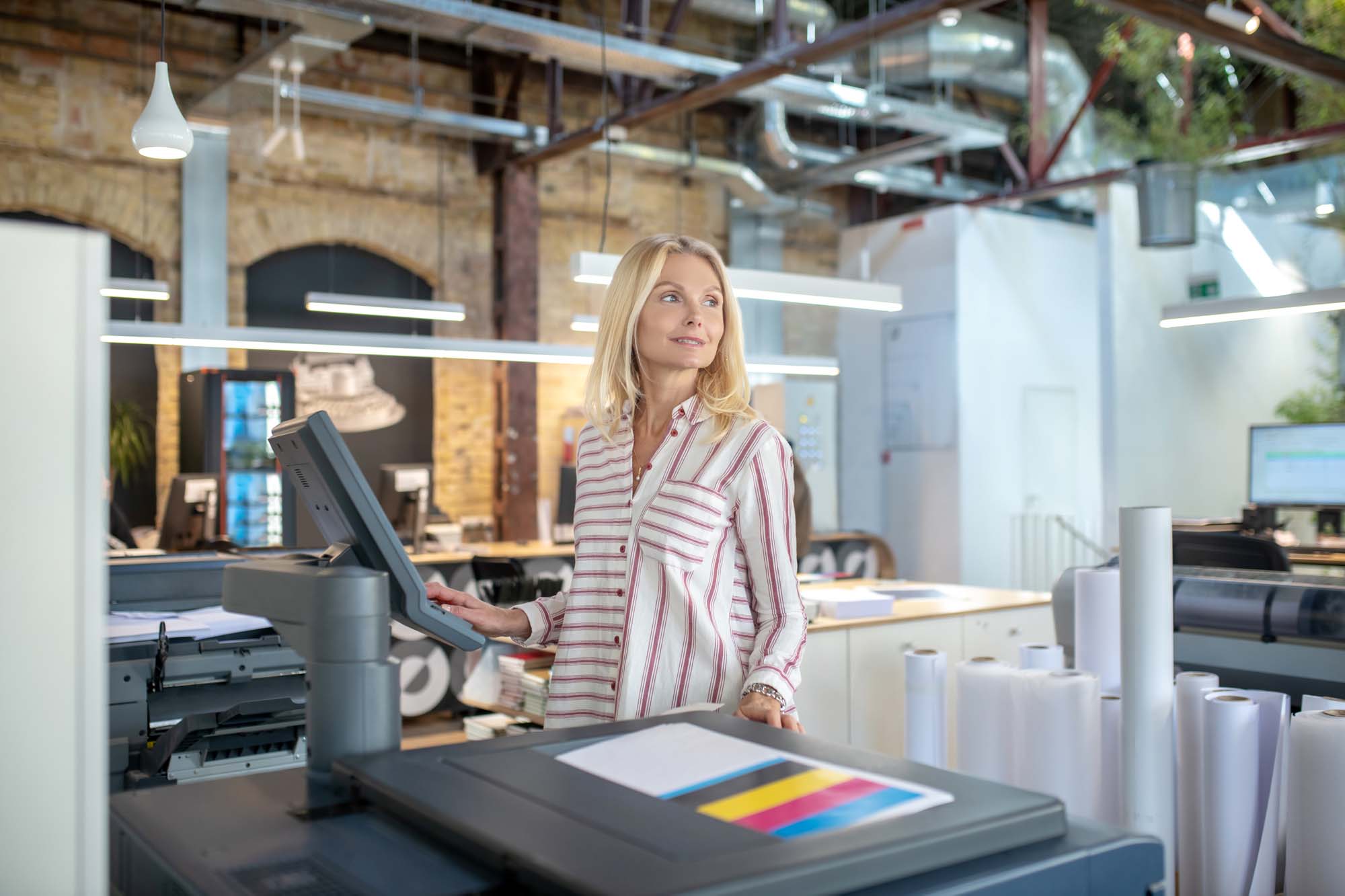 Print Tracking user interface showing a professional in a modern office using a large-format printer, with colorful print output and organized print supplies nearby.