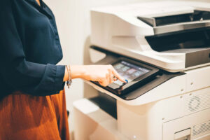 Office Printer Touchscreen – A woman operating a multifunction office printer using its touchscreen interface, wearing a navy blouse and orange pants.