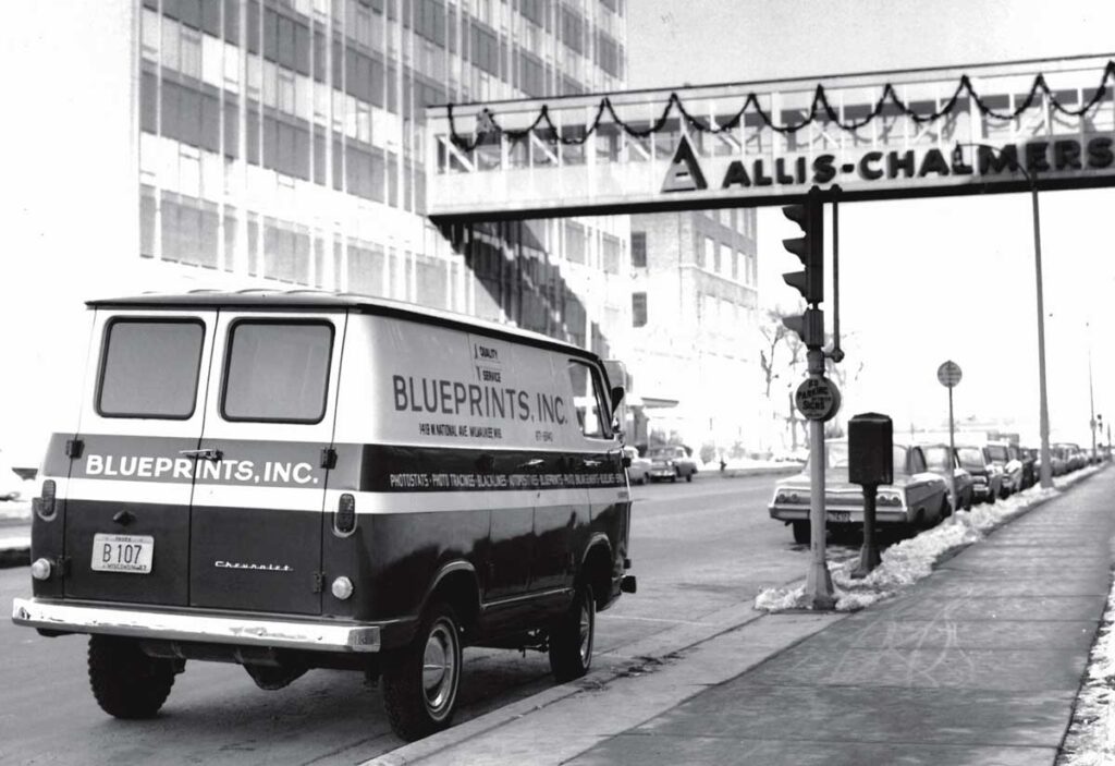 Archival black and white image of a Blueprints, Inc. delivery van parked near the Allis-Chalmers skywalk in Milwaukee during winter.