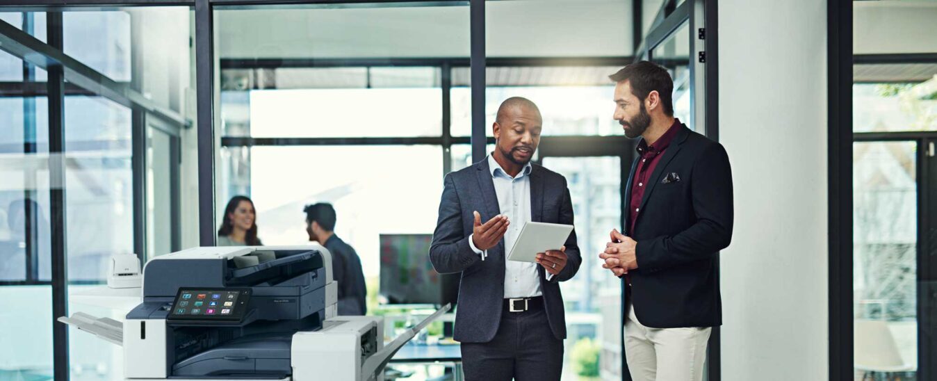 Printing Solutions Menu 3 Managed Print Services professionals discussing print strategy in a modern office setting, standing beside a Xerox multifunction printer with a tablet in hand.