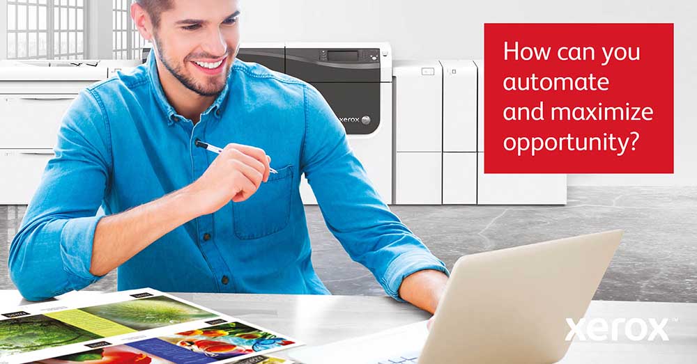 Xerox Print Automation – Smiling professional at a desk with a laptop and vibrant print samples, with a Xerox production printer in the background.