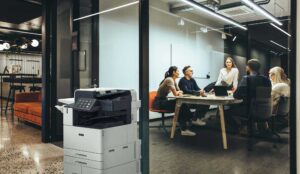 Xerox Lifestyle image featuring a modern office with a Xerox multifunction printer in the foreground and a team of professionals collaborating in a glass-walled meeting room.