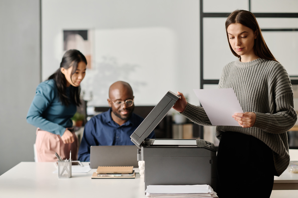 Office Printer being used by a woman in a gray sweater as two coworkers collaborate in the background at a modern, well-lit office workspace.