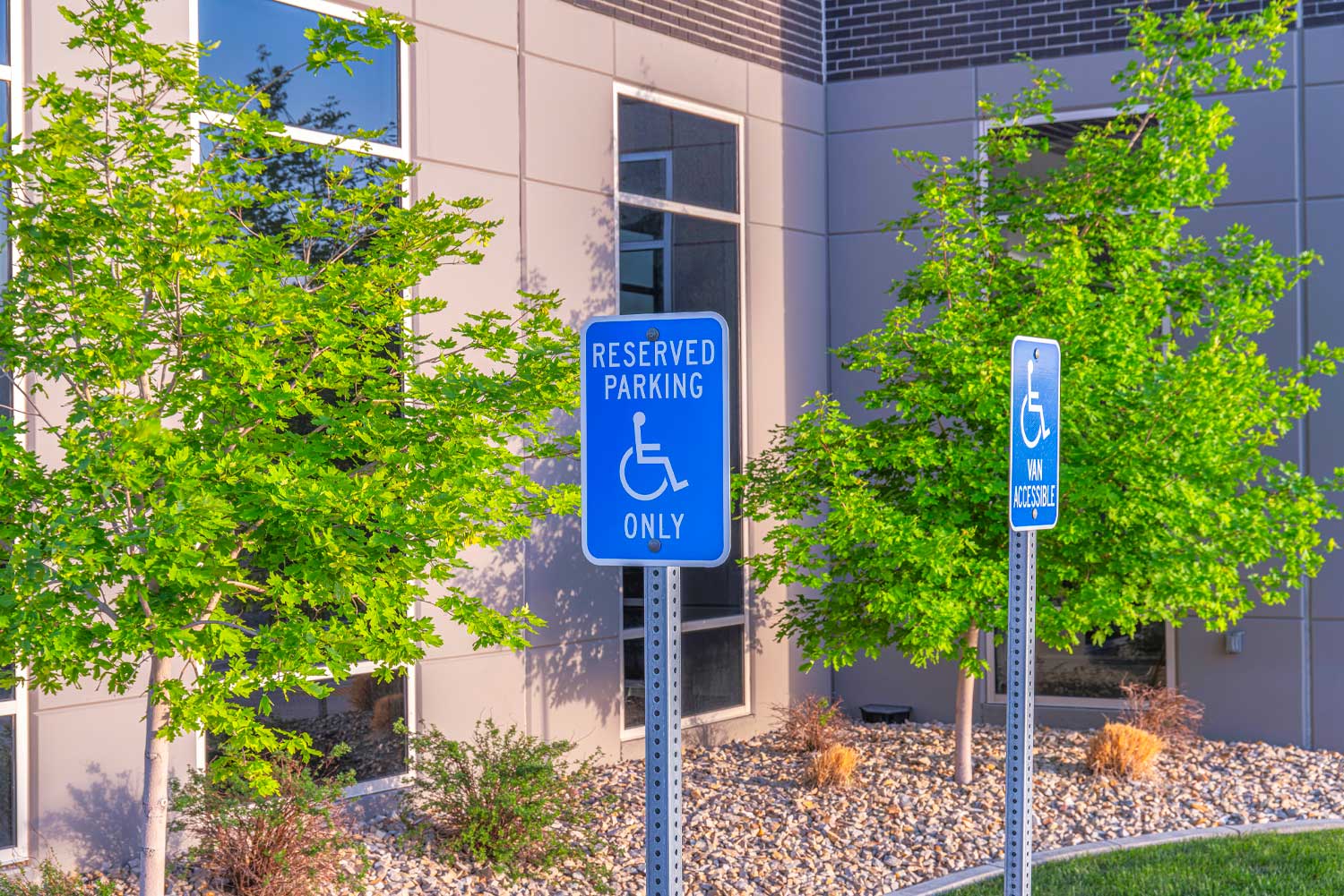 ADA Parking Lot Signage featuring blue reserved parking and van accessible signs outside a modern building with trees and landscaping in the background.