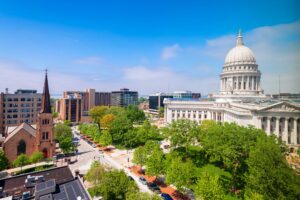 Madison Wisconsin skyline featuring the Wisconsin State Capitol, lush green trees, historic church, and surrounding downtown buildings under a clear blue sky.