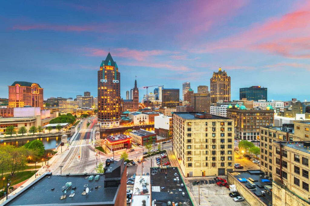 Milwaukee WI skyline at dusk showcasing downtown buildings, bridges, and city lights beneath a vibrant pink and blue sky.