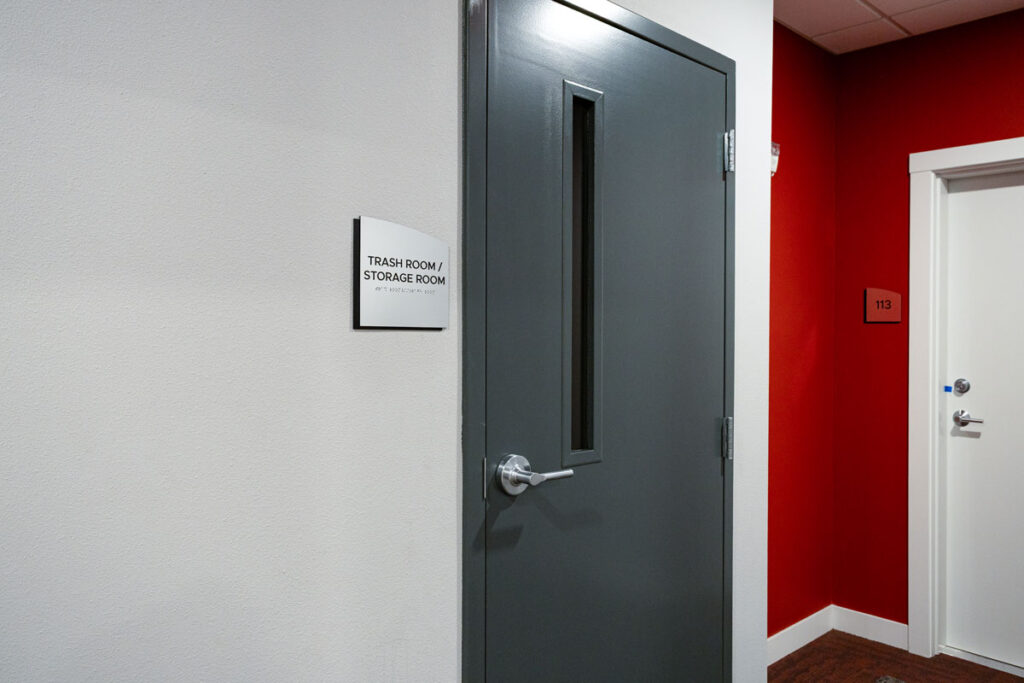 ADA Signage for trash and storage room at Colorado Commons features tactile lettering and braille beside a dark gray door in a modern residential hallway.