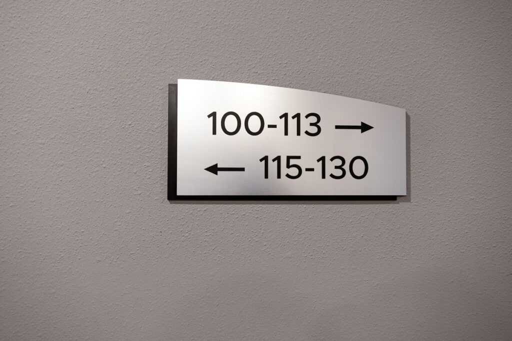 ADA Signage close-up of brushed metal room number directional sign showing arrows for rooms 100–113 and 115–130, mounted on a textured interior wall.