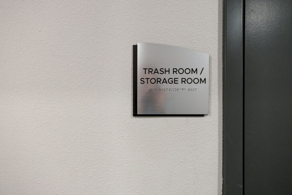 ADA Signage close-up of tactile braille sign labeled “Trash Room / Storage Room,” mounted beside a door on a white wall at Colorado Commons.