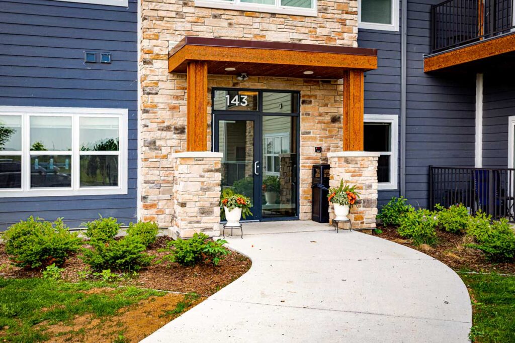 Colorado Commons front entrance view of building 143, featuring stone and wood architectural details, glass doors, potted plants, and a landscaped walkway.