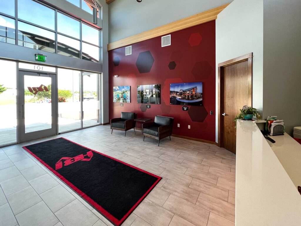 HBI Adhesive Wall Wrap in a bright, modern lobby featuring hexagon designs, mounted project photos, branded floor mat, and natural wood and glass accents.