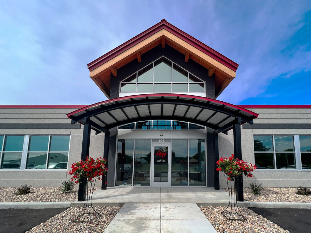 HBI front entrance featuring modern architecture with a covered canopy, red trim, and symmetrical stone landscaping on a clear blue-sky day.