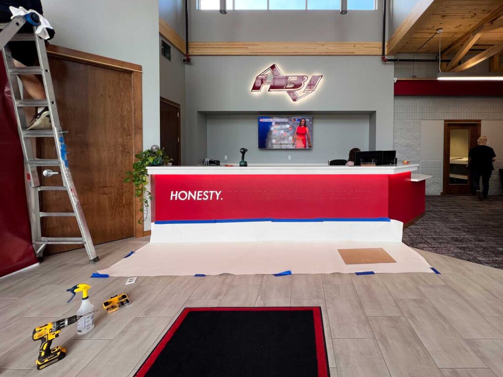 HBI Dimensional Signage Installation above a red-and-white reception desk, featuring the word “HONESTY” and tools visible during final setup stages.