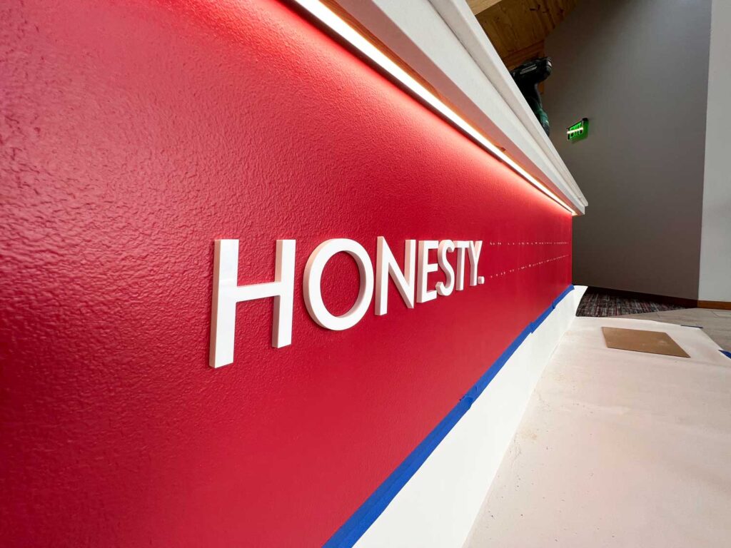 HBI Dimensional Lettering Installation close-up of the word “HONESTY” in raised white letters on a red reception desk with alignment guides visible.