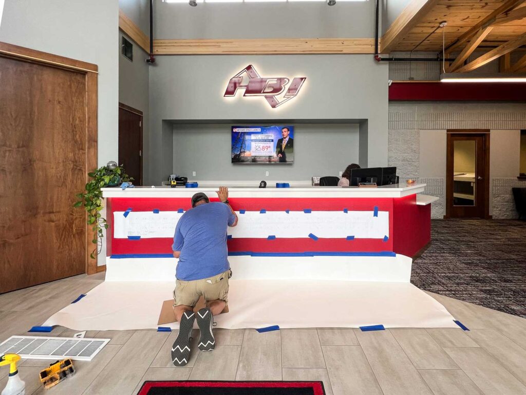 HBI Dimensional Lettering Install showing a technician applying branded vinyl lettering on the front of a red-and-white reception desk with prep materials on the floor.