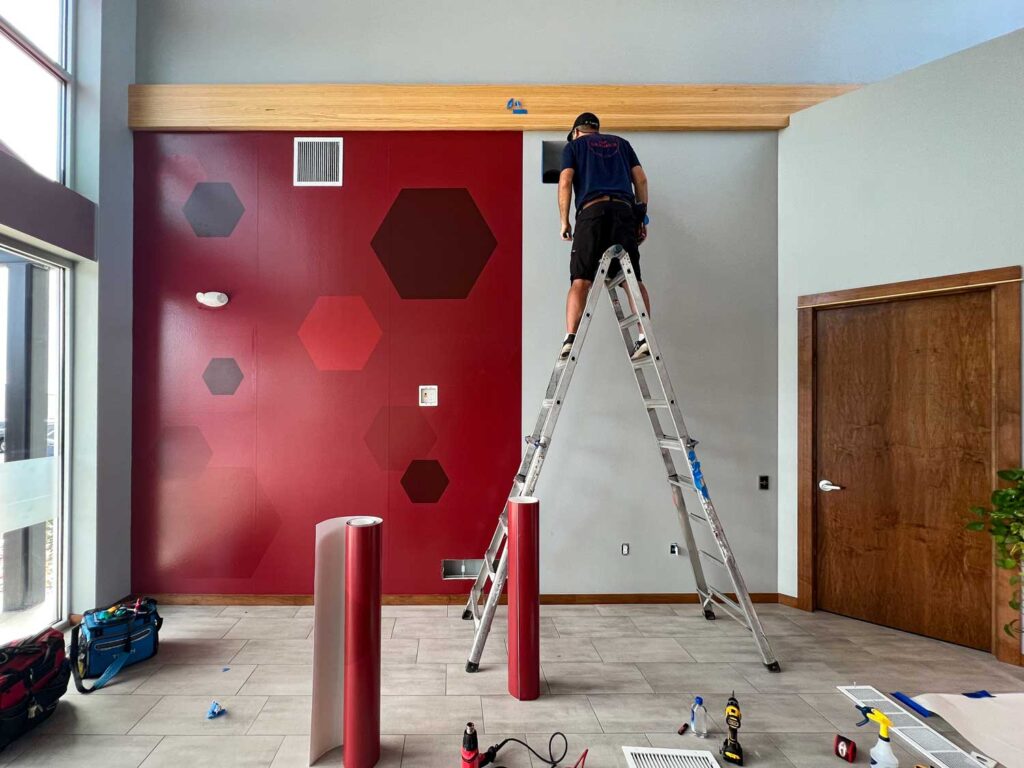 HBI Adhesive Wall Graphics Installation in progress as a technician applies red and dark hexagonal wall graphics from a ladder in a modern lobby interior.