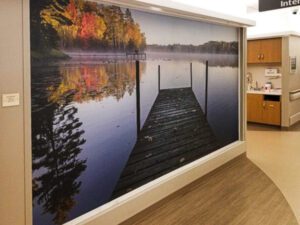 Adhesive Wall Art mural in a healthcare hallway featuring a peaceful lake with a dock, surrounded by autumn trees and a soft morning mist.