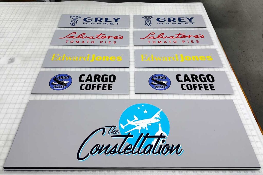 Branded Environmental Signage displayed on a table, showcasing nameplates for businesses like Grey Market, Salvatore's Tomato Pies, Edward Jones, Cargo Coffee, and The Constellation, all designed with distinct colors and logos.