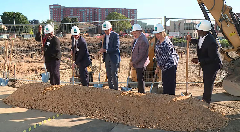 Triangle Neighborhood Ground Breaking ceremony featuring six individuals in hard hats using shovels to break ground at a construction site with urban backdrop.