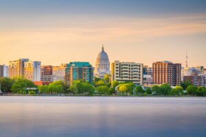 Madison Wisconsin skyline at sunset with the Wisconsin State Capitol building centered among modern buildings, viewed across a calm lake with green trees in the foreground.